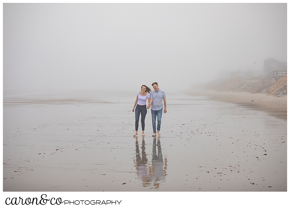 man and woman walking in the fog, on the beach, holding hands during a dreamy maine engagement session